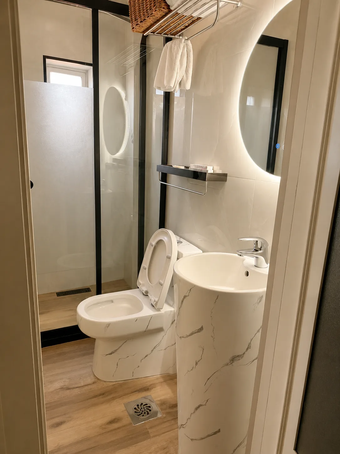 Casa de banho em mármore branco com espelho redondo iluminado, lavatório coluna, sabonetes na prateleira, toalhas penduradas e cabina de duche com base em madeira