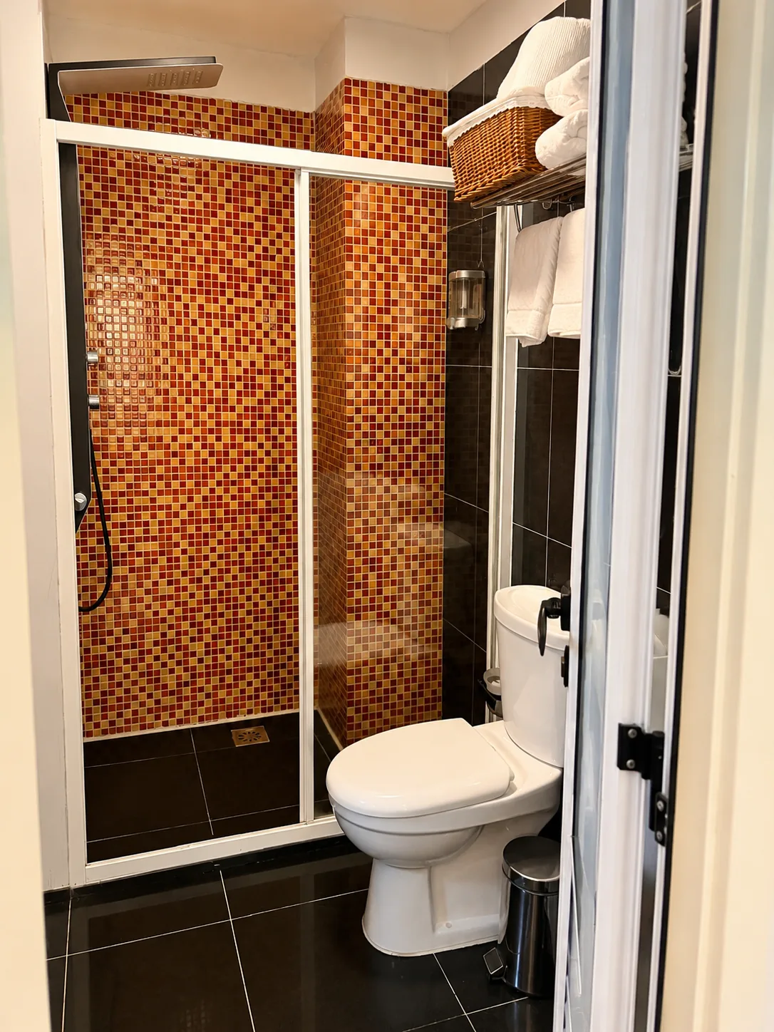Casa de banho privativa com cabina de duche de portas de correr, parede em mosaico vermelho e dourado, chuveiro de tecto e de mão, sanita e toalhas brancas dobradas num cesto de verga
