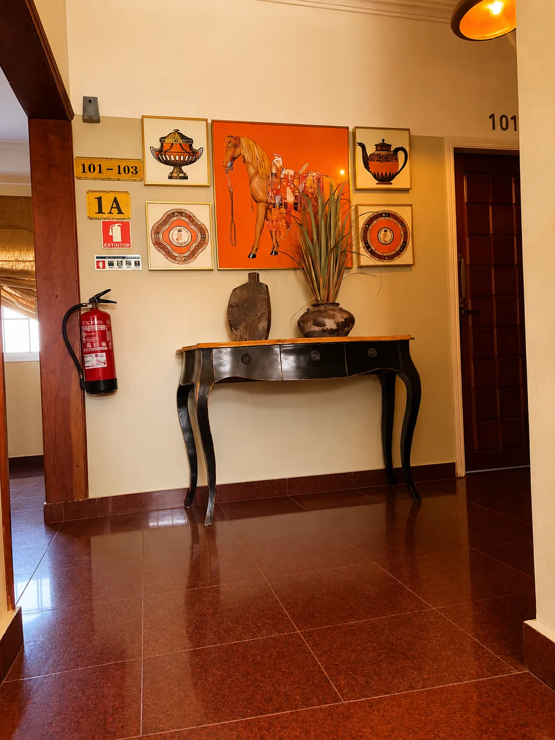 Corredor do primeiro piso com placa dos quartos 101-103, consola preta de pernas curvas, jarra e quadros emoldurados, extintor na parede e chão de pedra polida à porta do quarto 101