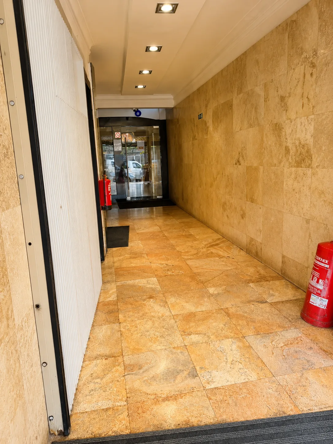 Entrada coberta da residencial, com chão e paredes em mármore claro, tecto com focos de luz, extintor na parede e porta de vidro para a recepção