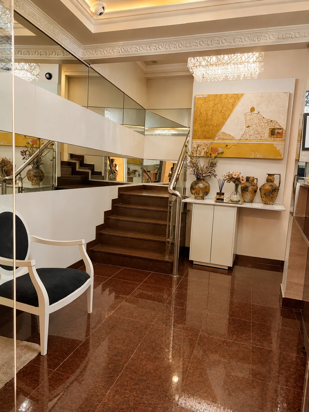 Escadaria junto à recepção, com degraus em pedra, corrimão de inox, parede de espelhos, cadeira estofada escura, consola com jarras e câmara de videovigilância no tecto