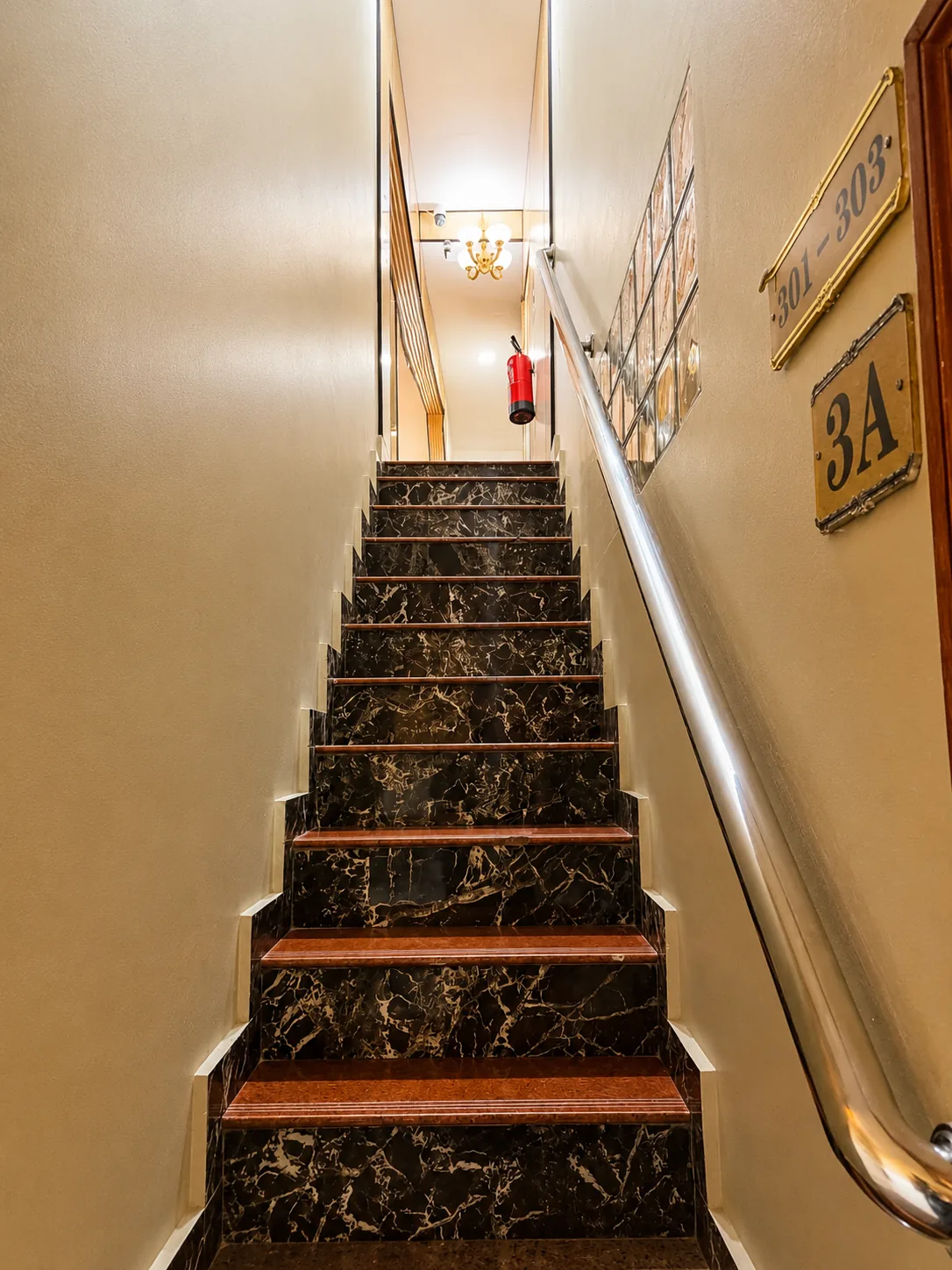 Escadas para o terceiro piso, com espelhos dos degraus em mármore escuro e pisos em madeira, corrimão de inox, placas dos quartos 301-303 e extintor no patamar