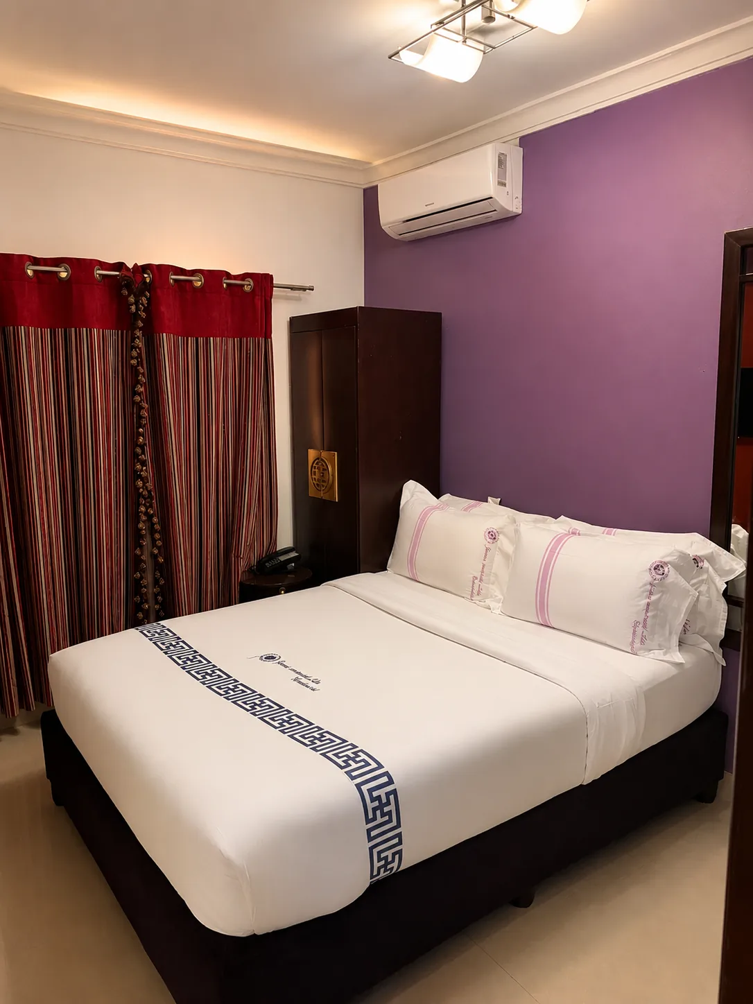 Quarto Casal com cama de casal, parede de destaque roxa, ar condicionado, roupeiro de madeira escura e cortinados às riscas