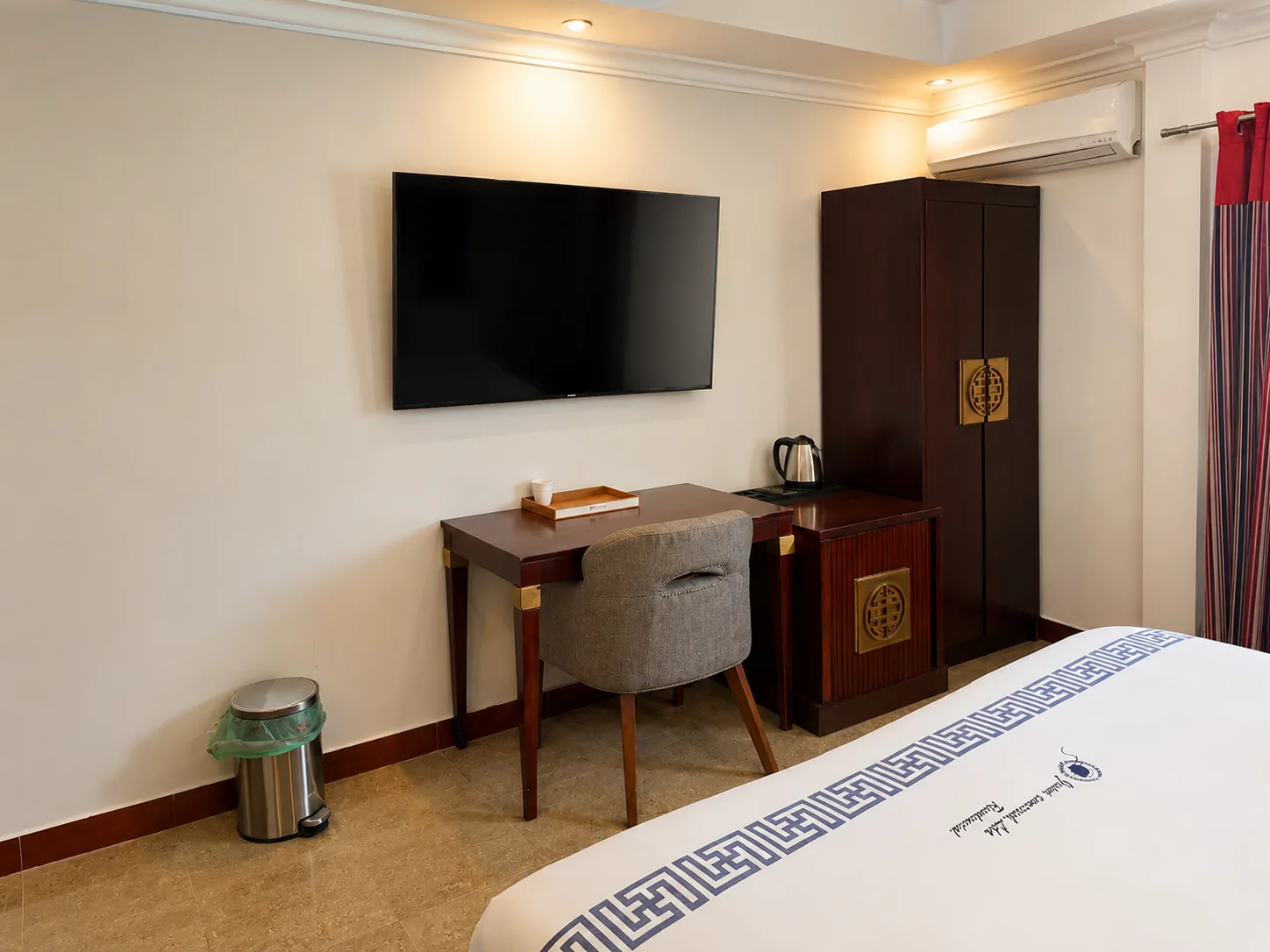 Quarto com televisão de ecrã plano na parede, secretária de madeira com cadeira estofada, jarro eléctrico, roupeiro alto, ar condicionado e cama com colcha branca