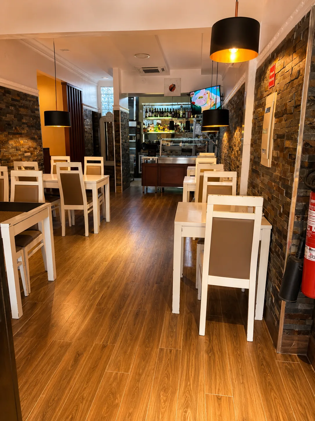 Sala de restaurante com mesas e cadeiras brancas, chão de madeira, paredes em pedra e o balcão do bar ao fundo